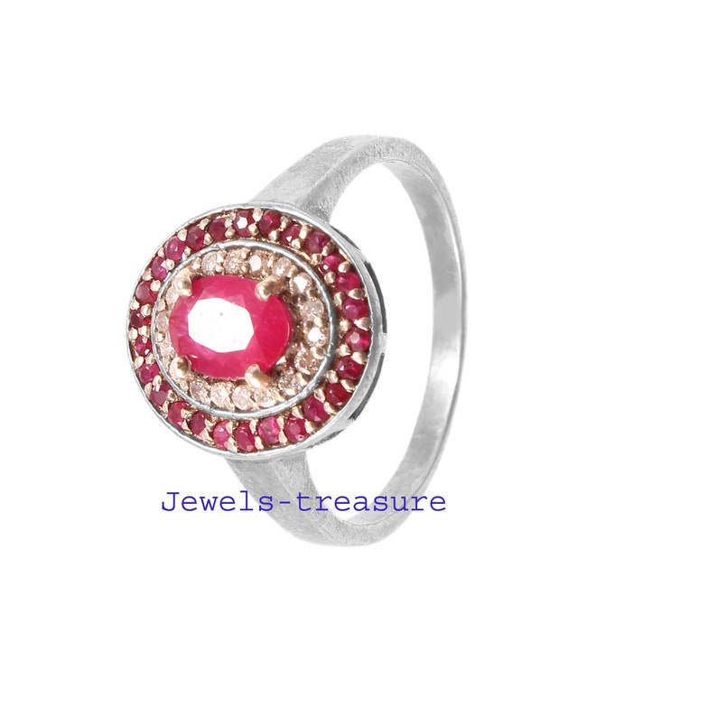 Op de afbeelding: Een zilveren ring met een grote ovale robijn als middensteen, omringd door een halo van kleinere rode edelstenen en een ring van heldere stenen. De ring heeft een eenvoudige, gepolijste zilveren band. De tekst "Jewels-treasure" staat onderaan.
