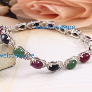 May include: A silver-toned bracelet featuring a series of alternating oval-shaped cabochon gemstones in red, blue, and green. The stones are set in metallic prongs and connected by small silver links. The text Jewelstreasure is visible on the image.