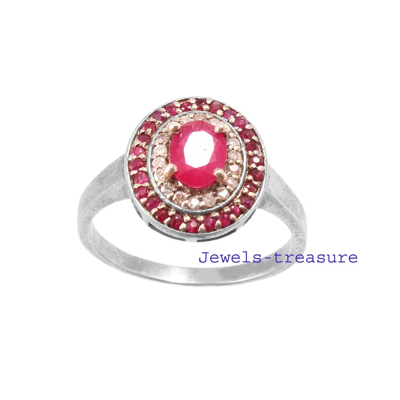 Op de afbeelding: Een zilveren ring met een grote, ovale robijn als middensteen, omringd door een halo van kleinere rode edelstenen en een ring van kleinere, heldere stenen. De ring heeft een eenvoudige band en de tekst "Jewels-treasure".