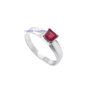 Pode incluir: Um anel de tom prateado com uma pedra preciosa de corte quadrado vermelho profundo num engaste de quatro garras. A banda é lisa e polida com uma largura uniforme. O texto 'jewels-treasure' é visível em azul perto da pedra.