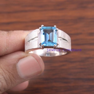 May include: A silver ring featuring a rectangular emerald-cut light blue gemstone set in the center. The band has a textured, cross-hatched finish on the sides with smooth silver accents. The text jewels-treasure is visible on the band.