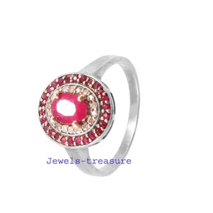 Op de afbeelding: Zilveren ring met een ovale, rode middensteen, omringd door kleinere rode en witte edelstenen. De ring heeft de tekst "Jewels-treasure" op de band.