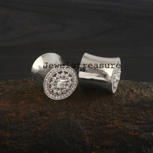 May include: A pair of silver-toned ear plugs with a flared cylindrical shape. The front face of each plug features a circular arrangement of small clear stones surrounding a central square stone, with the text Jeweltreasure overlaid.