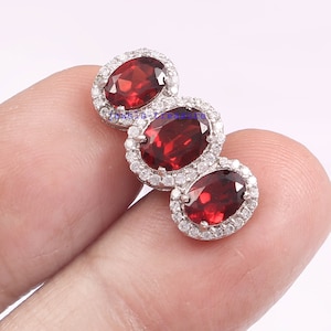 May include: Three oval-shaped red gemstones set in silver with a halo of smaller clear gemstones.