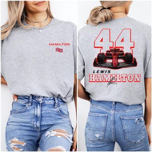 K&ouml;nnte beinhalten: Graues T-Shirt mit "HAMILTON" und einer britischen Flagge auf der linken Seite. Die R&uuml;ckseite des Shirts zeigt ein rotes Rennwagen mit der Nummer "44" und "LEWIS HAMILTON" in roter Schrift.
