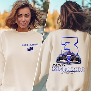May include: A cream-colored sweatshirt with a blue number "3" on the back, a Formula 1 car, and the text "Daniel Ricciardo" with a signature. The front of the sweatshirt has the text "Ricciardo" and an Australian flag.