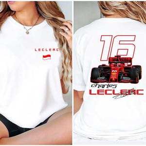 May include: White t-shirt with a red and white flag and the text "Leclerc" on the front. The back of the shirt has a red race car with the number "16" and the text "Charles Leclerc" in red.