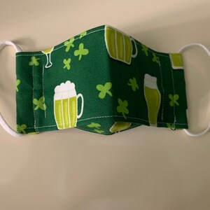May include: A green face mask with a white elastic band. The mask has a pattern of green shamrocks and beer mugs.