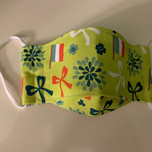 May include: A green fabric face mask with a white elastic band. The mask has a pattern of white bows, green flowers, and red and white flags.