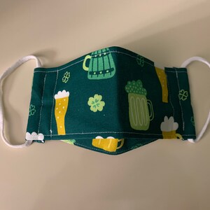 May include: A green fabric face mask with a white elastic band. The mask has a repeating pattern of beer mugs and shamrocks.