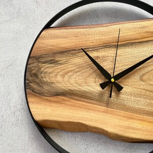 May include: A wooden wall clock with a black metal frame and black hands. The clock face is made of a natural wood slice with a unique grain pattern.