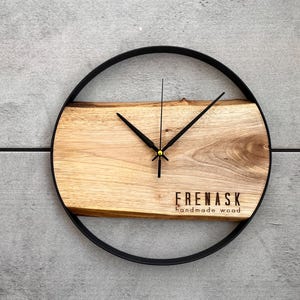 May include: A round wooden clock with black hands and a black metal frame. The clock face is made of a natural wood slice with a unique grain pattern. The clock face also has the text "FRENASk handmade wood" printed on it.