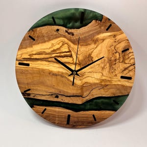 May include: A round wooden wall clock with a green epoxy resin inlay. The clock face has black hour and minute hands and black Roman numeral markings.