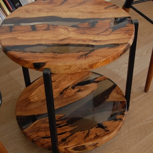 May include: A two-tiered side table with a round wooden top and bottom. The top is made of wood with a clear epoxy resin fill, creating a unique pattern. The table has a black metal frame.