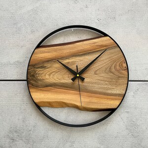 May include: A round wooden clock with a black metal frame. The clock face is made of a natural wood slice with a unique grain pattern. The clock hands are black and the center of the clock has a gold accent.