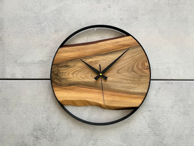 May include: A round wooden clock with a black metal frame. The clock face is made of a natural wood slice with a unique grain pattern. The clock hands are black and the center of the clock has a gold accent.
