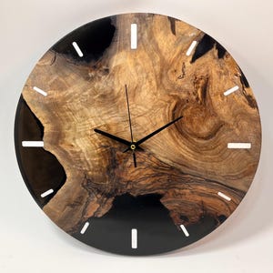 Könnte beinhalten: Eine runde Wanduhr aus Holz mit schwarzem Epoxidharz und weißen Stundenmarkierungen. Das Zifferblatt der Uhr besteht aus einer natürlichen Holzscheibe mit einem einzigartigen Maserungsmuster.