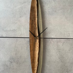 May include: A wooden and clear resin wall clock with black hands and a gold center. The clock is shaped like an oval and has a natural wood grain pattern.