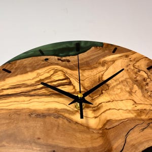 May include: A round wooden clock with black hands and a green epoxy resin inlay. The clock face is made of olive wood with a natural grain pattern.