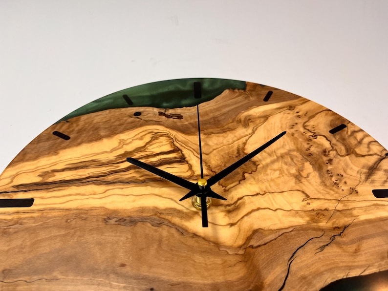 May include: A round wooden clock with black hands and a green epoxy resin inlay. The clock face is made of olive wood with a natural grain pattern.
