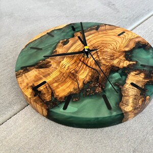 May include: A round wooden clock with a green resin inlay. The clock face has black hands and black Roman numeral markings.