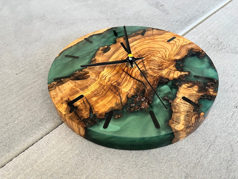 May include: A round wooden clock with a green resin inlay. The clock face has black hands and black Roman numeral markings.