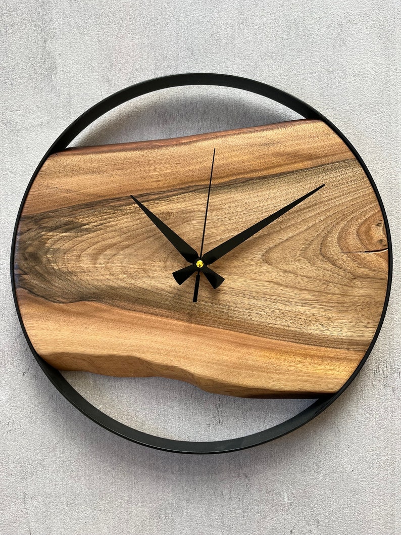 May include: A round wooden clock with a black metal frame. The clock face is made of a natural wood slice with a unique grain pattern. The clock hands are black and the center of the clock is gold.