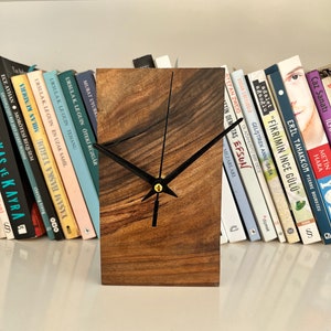 Op de afbeelding: Een houten klok met zwarte wijzers en een gouden centrum. De klok staat op een plank met een rij boeken erachter.
