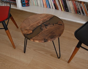 Op de afbeelding: Een ronde houten bijzettafel met een donkerbruin epoxyhars rivierontwerp. De tafel heeft een natuurlijke houtnerf en wordt ondersteund door drie zwarte haarspeldpoten. De tafel heeft een diameter van ongeveer 46 cm.