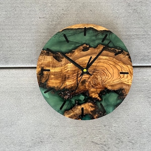 May include: A round wooden clock with a green epoxy resin inlay. The clock face has black hands and black Roman numeral markings.