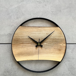 May include: A round wooden clock with black hands and a black metal frame. The clock face is made of a light brown wood with a darker brown wood grain.