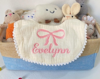 以下が含まれることがあります： ぬいぐるみとよだれかけが入ったベビーギフトバスケット。クリーム色のよだれかけにはピンクのリボンと「Evelynn」という名前が刺繍されています。バスケットはベージュとブルーで、ウサギのぬいぐるみと雲が入っています。