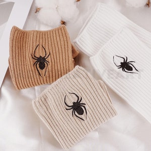 Puede incluir: Tres pares de calcetines con un diseño de araña. Un par es marrón, otro beige y otro blanco.