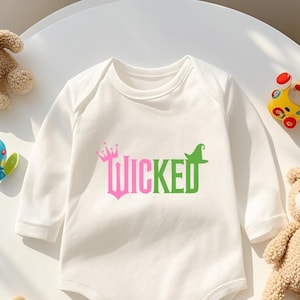 May include: White long-sleeved baby bodysuit with the word "WICKED" in pink and green, featuring a crown and witch hat design. The bodysuit is surrounded by teddy bears and toys.