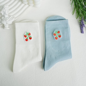 以下が含まれることがあります： 白い靴下と水色の靴下のペアで、それぞれに緑の葉が付いた赤いイチゴの刺繍が施されています。 靴下は柔らかい素材でできており、きれいに折りたたまれています。
