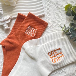 以下が含まれることがあります： オレンジと白の靴下1足。オレンジの靴下には「BIG BRO」と刺繍され、白い靴下には「lil sis」と刺繍されています。