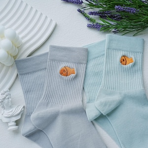 Pode incluir: Três pares de meias em tons de cinza e azul, cada um com um desenho de peixe laranja bordado. As meias são caneladas e têm um punho na parte superior. O fundo inclui elementos decorativos e raminhos de lavanda.