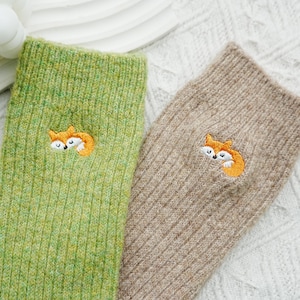 May include: Two cozy socks, one light green and one tan, are displayed. Each sock features an embroidered orange and white fox design. The socks are made of a soft, textured material, perfect for keeping feet warm.