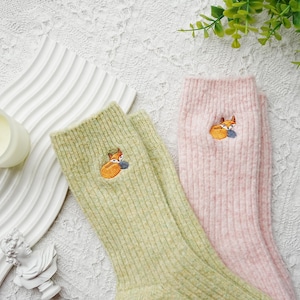 May include: A pair of ribbed knit socks, one in light green and the other in pink. Each sock features an embroidered orange fox with a blue ball. The socks are displayed on a white textured surface.