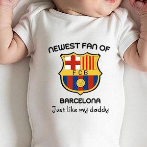May include: White baby onesie with the text "NEWEST FAN OF" and a soccer team logo with the text "FCB BARCELONA" and the text "Just like my daddy".