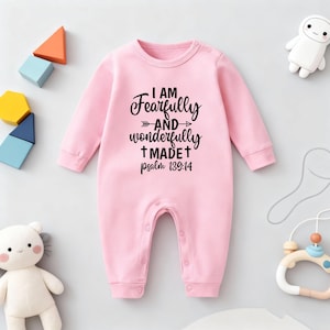 May include: A pale pink baby romper with long sleeves and popper fastenings. The romper displays the text "I AM Fearfully and wonderfully MADE psalm 139:14" in black script. Baby toys are scattered around the romper.