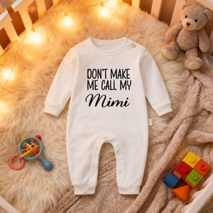 May include: A white baby romper with long sleeves and popper fastenings. The black text reads "DON'T MAKE ME CALL MY Mimi". A teddy bear and baby toys are in the background.