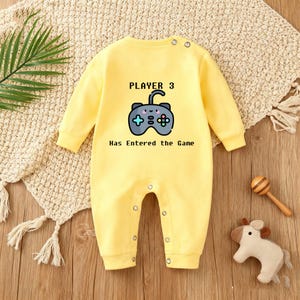 May include: A yellow baby onesie with long sleeves and snap closures. The front features a graphic of a video game controller and the text "PLAYER 3 Has Entered the Game". The onesie is made of a soft material.