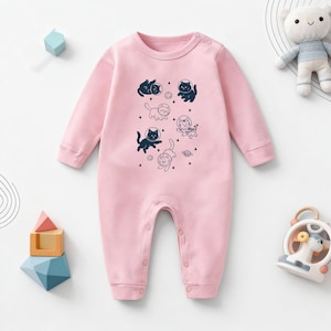 Può includere: Tutina rosa chiaro per neonati, con maniche lunghe e chiusure a scatto. La tutina presenta una stampa di gatti dei cartoni animati con caschi da astronauta e fluttuanti nello spazio. Il design include stelle e un pianeta.