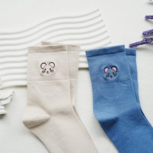 May include: Two pairs of socks, one beige and one blue, with embroidered mouse faces on the cuffs. The socks are made of a soft material, suitable for everyday wear. The mouse embroidery includes black eyes and whiskers.