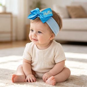 May include: A light blue fabric headband featuring a large, matching blue bow on the side. The bow is embellished with the name Hollie written in gold cursive script. The headband is made of a soft, stretchy material designed to fit comfortably.