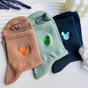 Op de afbeelding: Drie paar sokken met geborduurde Mickey Mouse-koppen in verschillende kleuren: oranje, groen en blauw. De sokken zijn bruin, groen en zwart.