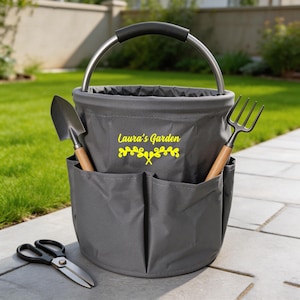 May include: A gray garden tool tote with a metal handle and pockets. The tote contains a trowel, a garden fork, and a pair of scissors. The text "Laura's Garden" is printed in yellow on the front.