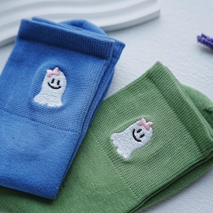 May include: Two pairs of ankle socks, one blue and one green, are shown. Each sock has an embroidered white ghost with a pink bow and a smiling face. The socks are made of a soft, knitted material.