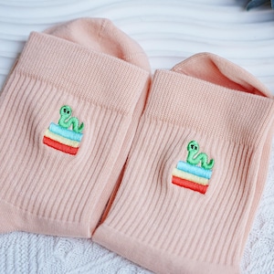 May include: A pair of light pink socks with a ribbed texture. Each sock features an embroidered design of a green worm on top of a stack of colorful stripes. The socks are made of a soft material.